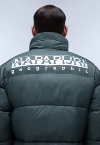 Giacca a vento verde con colletto alto. Presenta un grande logo bianco "NAPAPIJRI" e il testo "geographic" sulla schiena. Texture liscia e imbottita.
