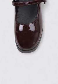 Zapato Mary Jane de charol burdeos con punta redonda y suela de goma negra, que presenta una sola correa en el empeine.