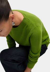 Grüner gestrickter Pullover mit rundem Halsausschnitt, strukturierten Details an den Schultern und gerippten Bündchen. Kombiniert mit dunkelblauen Jeans.