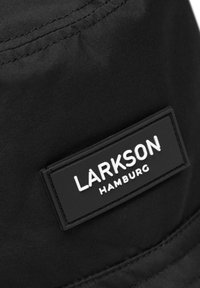 Musta kangaskorillinen hattu, jossa on suorakulmainen musta kumilappu, jossa on valkoiset kohopainetut tekstit "LARKSON HAMBURG" ja näkyvät ompeluyksityiskohdat.