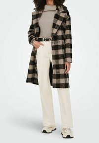 Manteau à carreaux noir et beige en laine, associé à un pull en maille beige, un pantalon blanc à jambes droites et des baskets légères.