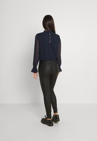 Navy blouse with textured fabric, ruffled collar, and button closure at the back. Paired with black fitted pants and black shoes.