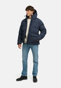 Giubbotto imbottito blu navy con chiusura a zip, cappuccio e due tasche frontali. Indossato sopra un maglione leggero e abbinato a jeans blu e stivali neri.