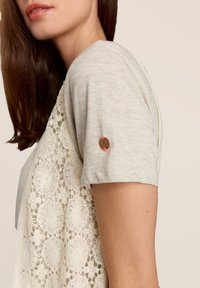 T-shirt gris clair à manches courtes, avec un dos en dentelle et un bouton décoratif avec un accent de fil rouge sur la manche.