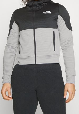 Osoba ubrana w czarno-szarą kurtkę North Face na suwak oraz czarne teksturowane spodnie, stojąca na jednolitym tle.