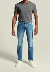 Mężczyzna w dopasowanej szarej koszulce, jasno-niebieskich dżinsach i białych sneakersach, stojący na tle jednolitego jasnego tła.