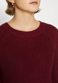 Maglione lavorato a maglia bordeaux con scollo rotondo e motivo a coste testurizzate, abbinato a orecchini a cerchio dorati e un accento di perline rosse.