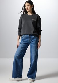 Sudadera gris oscuro con puños acanalados, combinada con jeans de pierna ancha azul. El conjunto se complementa con zapatillas blancas.
