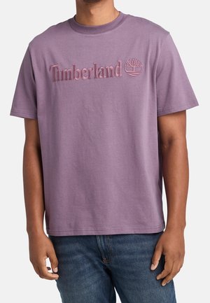 Mann trägt ein lila Timberland T-Shirt mit gesticktem Logo, kombiniert mit blauen Jeans, gezeigt von Hals bis Hüfte vor weißem Hintergrund.