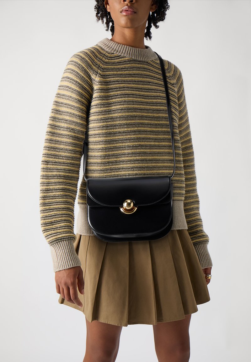 Borsa a spalla in pelle nera con pattina arrotondata, dettagli in hardware dorato e finitura liscia, abbinata a un maglione a righe in maglia e a una gonna plissettata.