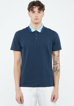 Giovane uomo con capelli ricci che indossa una polo blu scuro con colletto azzurro e pantaloni bianchi, in piedi davanti a uno sfondo semplice.