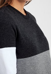 Mujer vistiendo un suéter con bloques de color en secciones negras, grises y blancas, mostrando un primer plano de la parte superior del torso y el cuello.