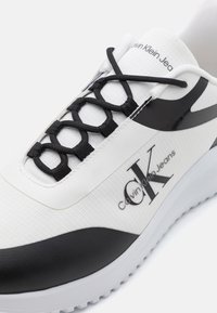 Valged spordijalatsid, millel on tekstureeritud pealispind, mustad aktsendid ja eristuv sidumissüsteem. Kaunistatud "Calvin Klein Jeans" logoga. Kummist tald.