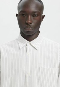 Stripete skjorte med knapper i lett stoff med tynne, vertikale linjer. Har spiss krage og brystlomme. Fargepaletten er hvit og lys grå.