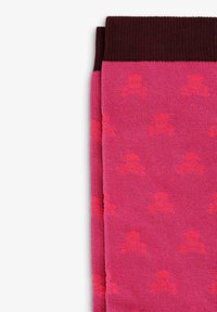 Calcetines rosas con un patrón de punto texturizado que presenta motivos de calaveras y tibias rojas. Puño acanalado marrón en la parte superior.