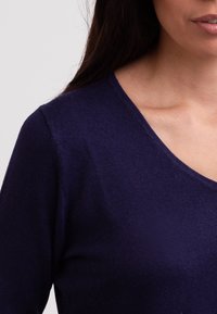 Tmavě fialový svetr s výstřihem do V, jemným žebrováním, měkkou texturou a přiléhavým střihem, předvádějící hladkou pletenou látku.