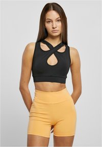 Crni crop top s prekrivenim trakama i dizajnom u obliku ključa, uparen s kratkim hlačama u breskvastoj boji visokog struka. Ukupno glatka tekstura.