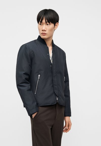 Νεαρός άνδρας ντυμένος με σκούρο μπλε μπουφάν bomber με ασημένιες τσέπες με φερμουάρ και σκούρο καφέ παντελόνι, στέκεται με το ένα χέρι στην τσέπη κοιτάζοντας προς τα αριστερά.