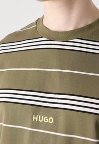 T-shirt en coton vert à manches courtes, présentant des rayures horizontales blanches et noires, avec "HUGO" brodé en jaune sur la poitrine.