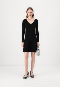 Black long-sleeve dress with a V-neckline, textured waist, and above-knee length. Paired with black heels and a silver handbag.