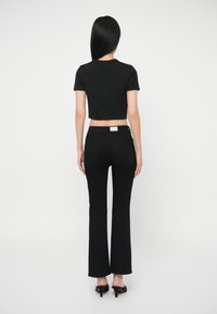 Γυναίκα με μακριά μαύρα μαλλιά στέκεται με την πλάτη γυρισμένη, φορώντας ένα μαύρο τοπ, φαρδιά μαύρα τζιν και μαύρες ψηλές παντόφλες σε λευκό φόντο.