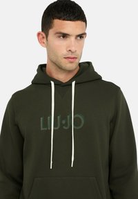 Donkergroene hoodie met een kangaroezak aan de voorkant, witte trekkoorden en een tonale logoprint. Zachte stof en een ontspannen pasvorm.