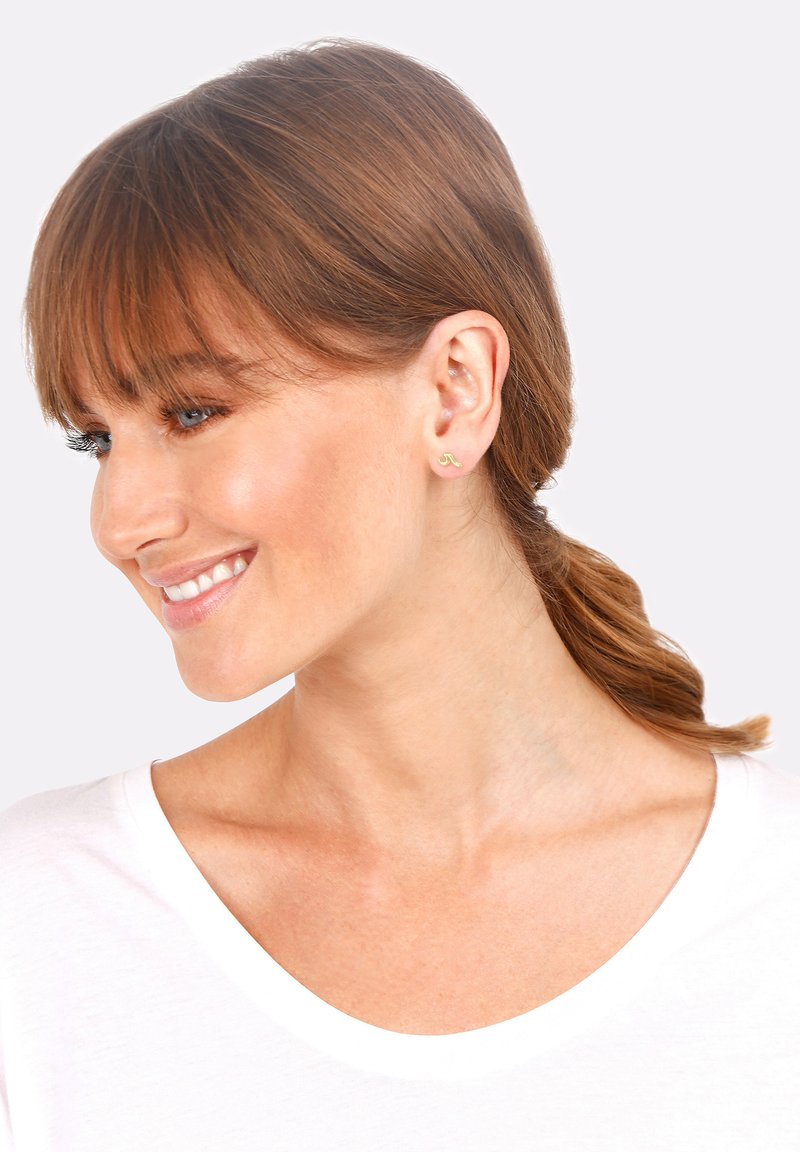 Gold earrings featuring a minimalist design in the shape of a small wave. The model has brown hair styled in a ponytail and wears a white shirt.