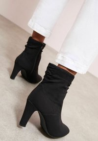 Botas negras de tobillo hechas de material suave, con un upper fruncido y cierre de cremallera, con un tacón alto y diseño de punta redonda.