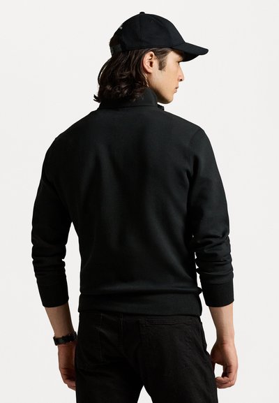 Zwarte sweatshirt met hoge kraag, geribbelde manchetten en een gladde textuur. Draag met zwarte broek en bijpassende pet, gezien vanachter.