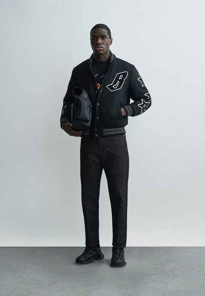 Νέος άνδρας που φοράει μαύρο varsity μπουφάν με λευκά κεντήματα, σκούρο τζιν, μαύρα αθλητικά παπούτσια, κρατώντας μαύρο σακίδιο πλάτης, στέκεται μπροστά σε μια απλή λευκή τοίχο.