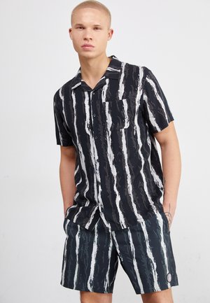 Camisa de botones de manga corta a rayas verticales en blanco y negro, con cuello y bolsillo. Combinada con pantalones cortos a juego con el mismo patrón.