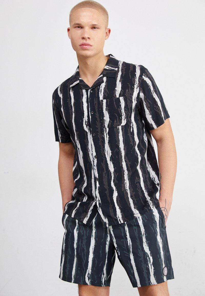 Camisa de botones de manga corta a rayas verticales en blanco y negro, con cuello y bolsillo. Combinada con pantalones cortos a juego con el mismo patrón.