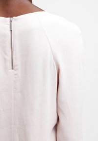 Top rosa chiaro realizzato in un tessuto liscio e strutturato, con chiusura a zip sul retro e design a scollo a barca, mostrato dalla vista posteriore.