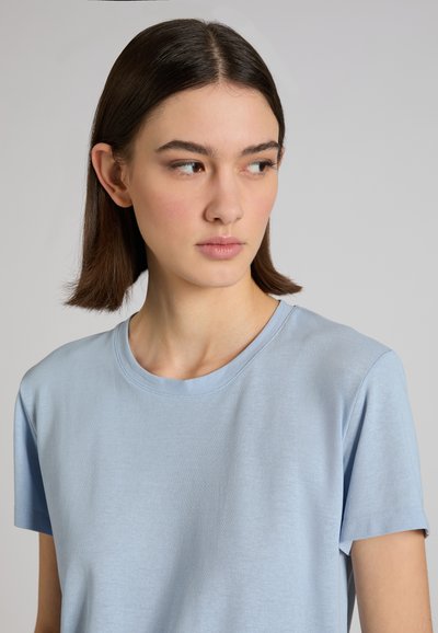 Jeune femme aux cheveux bruns raides portant une chemise à manches courtes bleu clair, regardant vers sa gauche sur un fond gris uni.