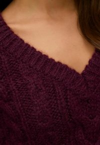 Pull en maille au col en V de couleur bourgogne profond, avec un motif de câble texturé et des bords côtelés. Matière douce et chaude en mélange de laine.