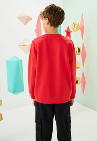 Jachetă roșie cu guler rotund, mâneci lungi și manșete strâmte, asortată cu pantaloni cargo negri. Fundalul prezintă decorațiuni geometrice.
