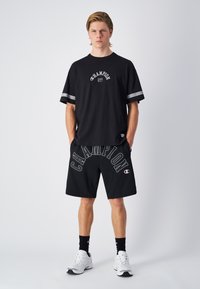 Camiseta de algodón negra con logo gris, combinada con pantalones cortos negros que presentan el texto blanco "CHAMPION". Incluidos zapatos deportivos blancos y calcetines negros.