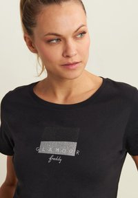 Camiseta de algodón negra con un diseño gráfico brillante que presenta un patrón rectangular y las palabras "GLAMOUR" y "freddy" en blanco. Cuello redondo.
