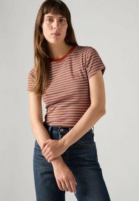 Mujer con cabello largo castaño, vistiendo una camiseta a rayas en tonos óxido, blanco y negro, y jeans azul oscuro, de pie con los brazos cruzados.