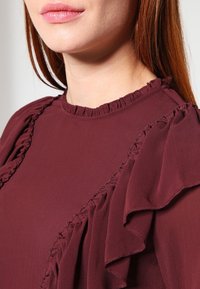 Femme portant un blouse bordeaux avec des détails en volants le long des épaules et de l'encolure, montrant son bas du visage et son cou.