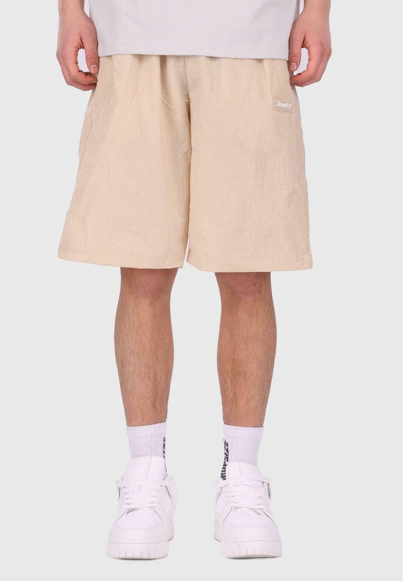 Shorts beiges avec un motif de grille texturé, taille élastique, poches latérales et petit logo blanc. Portés avec des baskets blanches et des chaussettes.