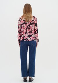 Donna con lunghi capelli castano chiaro, che indossa una blusa floreale rosa e nera, jeans blu e scarpe nere, girata di spalle su uno sfondo bianco.