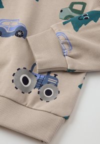 Beige pullover met blauw en groene voertuigpatronen, voorzien van ribgebreide manchetten en een zachte textuur. Opvallende details zijn een tractor en auto’s.