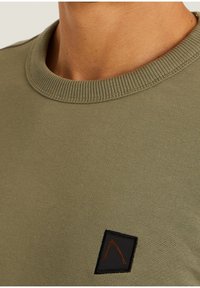 Sudadera de cuello redondo de algodón en color verde oliva con cuello acanalado. Presenta una etiqueta negra con un detalle del logo en naranja. Textura suave, diseño casual.