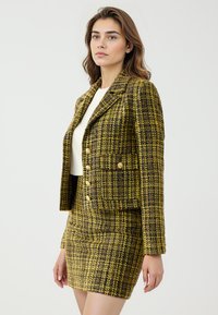 Tweed blazer in geel en zwart met een getailleerd ontwerp, dubbele rij knopen aan de voorkant, gouden knopen en een bijpassende rok. Glad, gestructureerd materiaal.