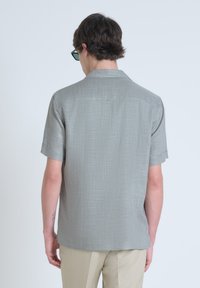 Camisa de manga corta con cuello en tejido gris claro, con un sutil patrón texturizado. Incluye un pequeño detalle de etiqueta en la parte posterior. Llevada con pantalones beige.