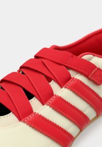 Κόκκινο αθλητικό παπούτσι με υφασμάτινες ρίγες σε κρεμ απόχρωση, κορδόνια από ύφασμα και επένδυση στο εσωτερικό σε κόκκινο χρώμα με μαλακά άκρα. Διακρίνονται λεπτομερείς ραφές.