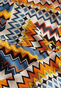 Tkanina z żywymi wzorami w zygzak i szewron w pomarańczowym, niebieskim, żółtym, czarnym i białym kolorze, pokazująca fałdy i teksturę.
