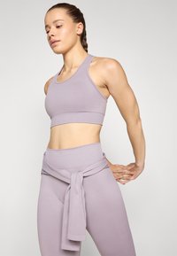 Soutien-gorge de sport lilas avec texture côtelée, associé à un legging taille haute assorti, présentant un détail en tissu noué autour de la taille.