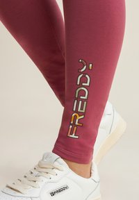 Leggings maroon con la grafica "FREDDY" in lettere colorate. Abbinati a sneaker bianche con logo e lacci testurizzati. Tessuto liscio.